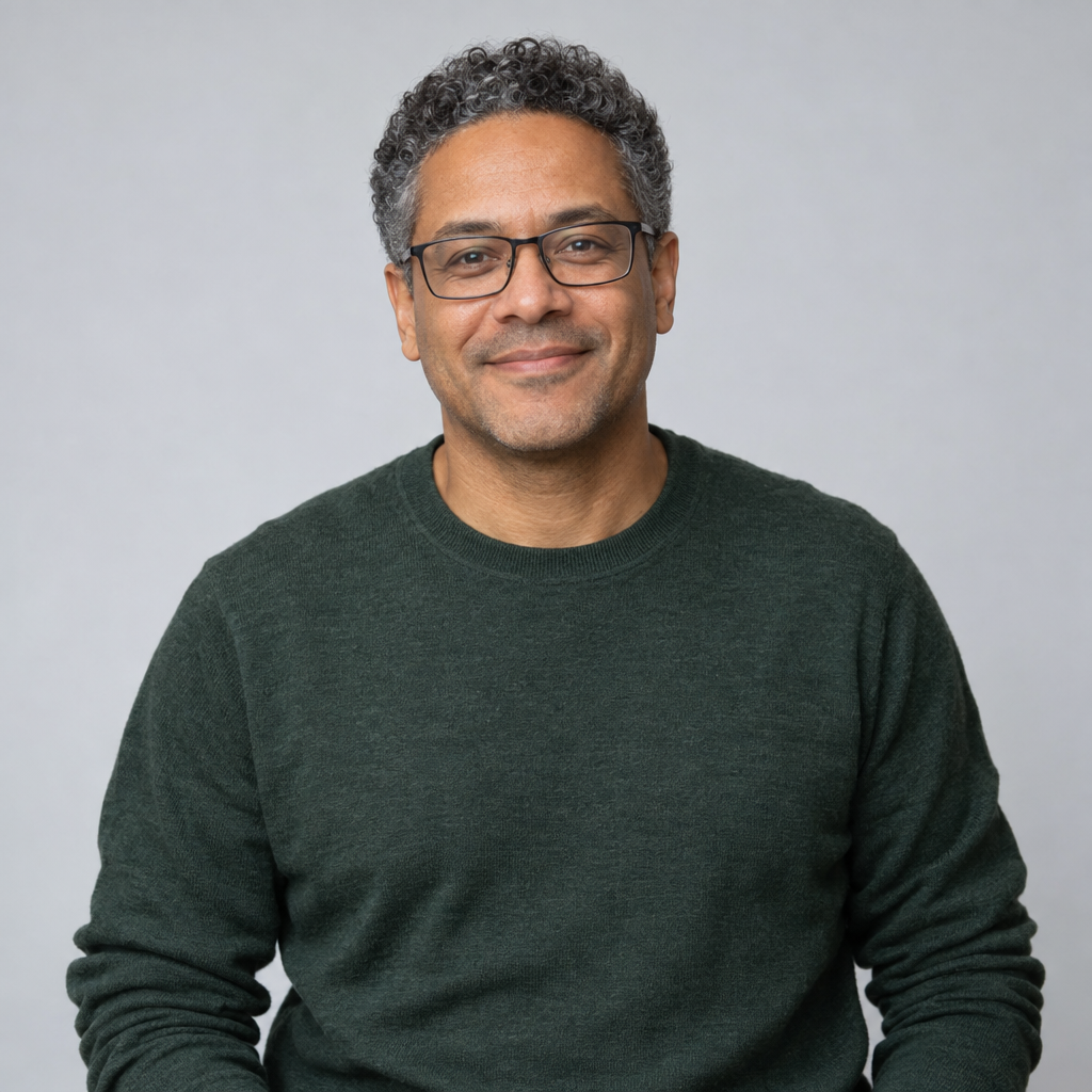 A centered, waist-up portrait of a smiling person with salt-and-pepper curls and rectangular glasses, wearing a dark green sweater against a pale-gray background.