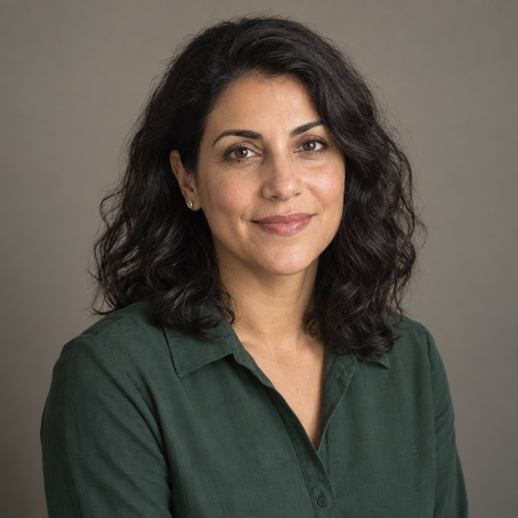A woman with voluminous wavy black hair and a calm, closed-mouth smile faces the camera in a dark green collared blouse, against a plain warm-gray background.